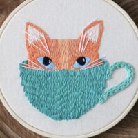 Delicately hand stitched ginger kitten poking its head out of a teal mug