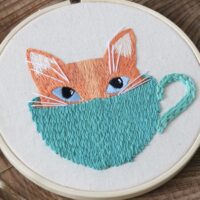 close up of hand stitches of a ginger kitten and a teal mug