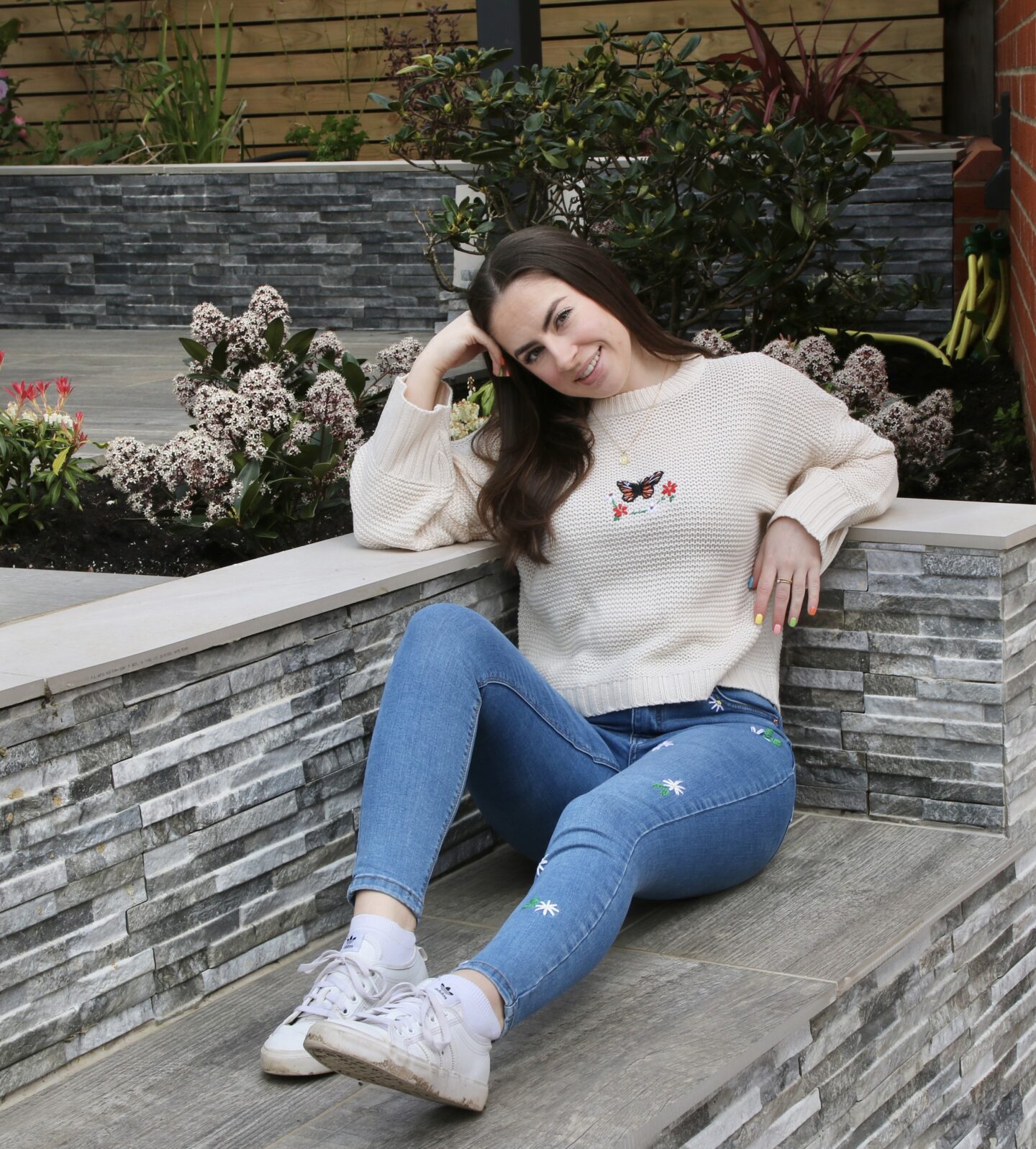 Brunette girl smiling and wearing blue skinny jeans hand embroidered with daisies and a white jumper hand embroidered with a butterfly