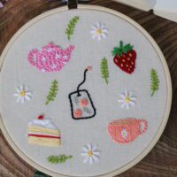 Hand embroidered afternoon tea themed pieces including: a pink teapot, pink teacup, strawberry, slice of cake, and rose teabag