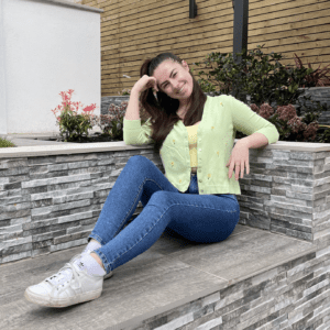 Brunette girl sitting on a bench wearing blue skinny jeans, a yellow top, and a green cardigan hand embroidered with sunflower