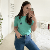 Brunette girl wearing a turquoise blue tee hand embroidered with a navy blue sailboat paired with blue jeans