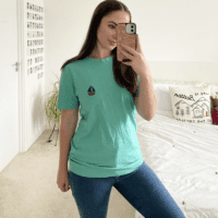 Brunette girl wearing a turquoise blue tee hand embroidered with a navy blue sailboat paired with blue jeans