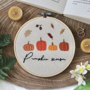 Embroidery hoop with “Pumpkin season” stitched in black thread, surrounded by embroidered pumpkins and autumn leaves on a cream fabric, placed on a wooden surface with decorative items.