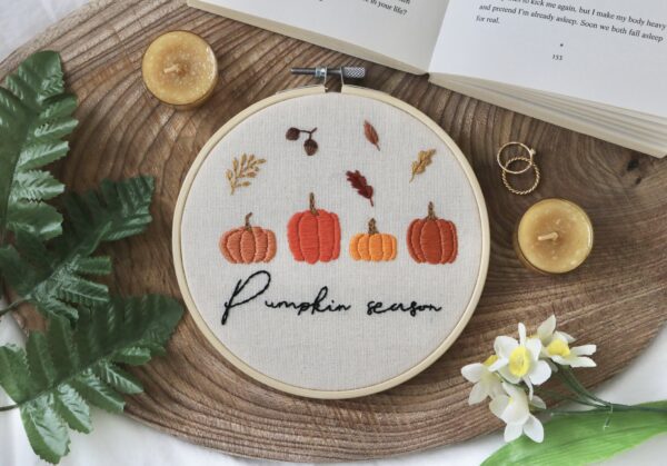 Embroidery hoop with “Pumpkin season” stitched in black thread, surrounded by embroidered pumpkins and autumn leaves on a cream fabric, placed on a wooden surface with decorative items.