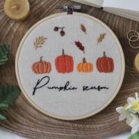 Embroidery hoop with “Pumpkin season” stitched in black thread, surrounded by embroidered pumpkins and autumn leaves on a cream fabric, placed on a wooden surface with decorative items.