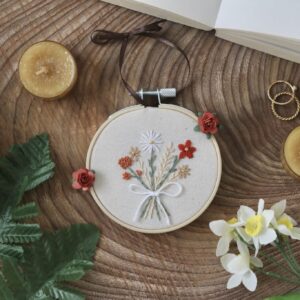 This image features a small, hand-embroidered hoop with a bouquet of flowers in autumnal colors, including white, orange, and beige, tied with a white ribbon. The embroidery is displayed in a wooden hoop, which is tied with a chocolate brown ribbon at the top, placed on a wooden surface.