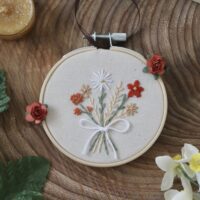 A small, hand-embroidered hoop featuring a bouquet of flowers in autumnal colors, including white, orange, and beige, tied with a white ribbon. The embroidery is displayed in a wooden hoop with red rose embellishments, placed on a wooden surface surrounded by a green leaf, yellow daffodils, golden candles, and gold rings.