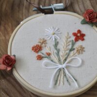 A small, hand-embroidered hoop featuring a delicate bouquet of flowers in autumnal colors, including white, orange, and beige blooms, tied together with a white ribbon. The embroidery is displayed in a wooden hoop, resting on a wooden surface with a red rose embellishment attached to the hoop.