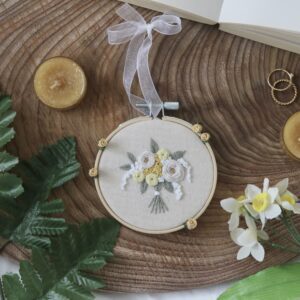 A flat lay image featuring a round embroidery hoop with a floral design on a wooden surface. The embroidery shows white and yellow flowers with green leaves, accented by small yellow roses. Hand tied with a white ribbon.