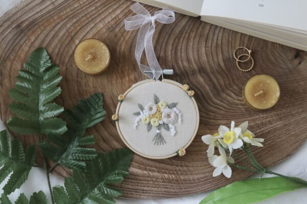 A flat lay image featuring a round embroidery hoop with a floral design on a wooden surface. The embroidery shows white and yellow flowers with green leaves, accented by small yellow roses. Hand tied with a white ribbon.