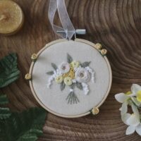 Embroidery hoop with a floral design featuring white and yellow roses on a beige fabric background, displayed on a wooden surface.