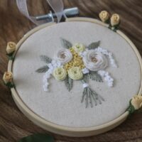 Embroidery hoop with a floral design featuring white and yellow roses on a beige fabric background, displayed on a wooden surface.