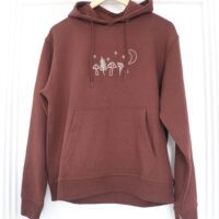 A dark chocolate brown coloured hoodie hanging on a wooden hanger, featuring hand-embroidered designs of mushrooms, leafy plants, stars, and a crescent moon across the chest. The hoodie has a front pocket and a small embroidered flower on the sleeve cuff