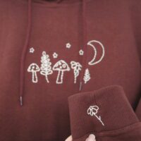 A dark chocolate brown coloured hoodie featuring hand-embroidered designs of mushrooms, leafy plants, stars, and a crescent moon across the chest. With a hand embroidered flower on the sleeve cuff