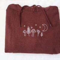 A dark chocolate brown coloured hoodie featuring hand-embroidered designs of mushrooms, leafy plants, stars, and a crescent moon across the chest.