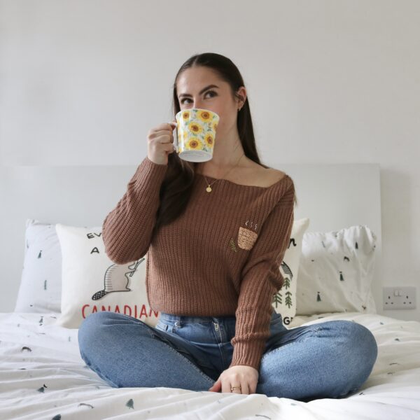 A brunette girl sat on a bed wearing blue jeans and a brown jumper hand embroidered with a coffee mug