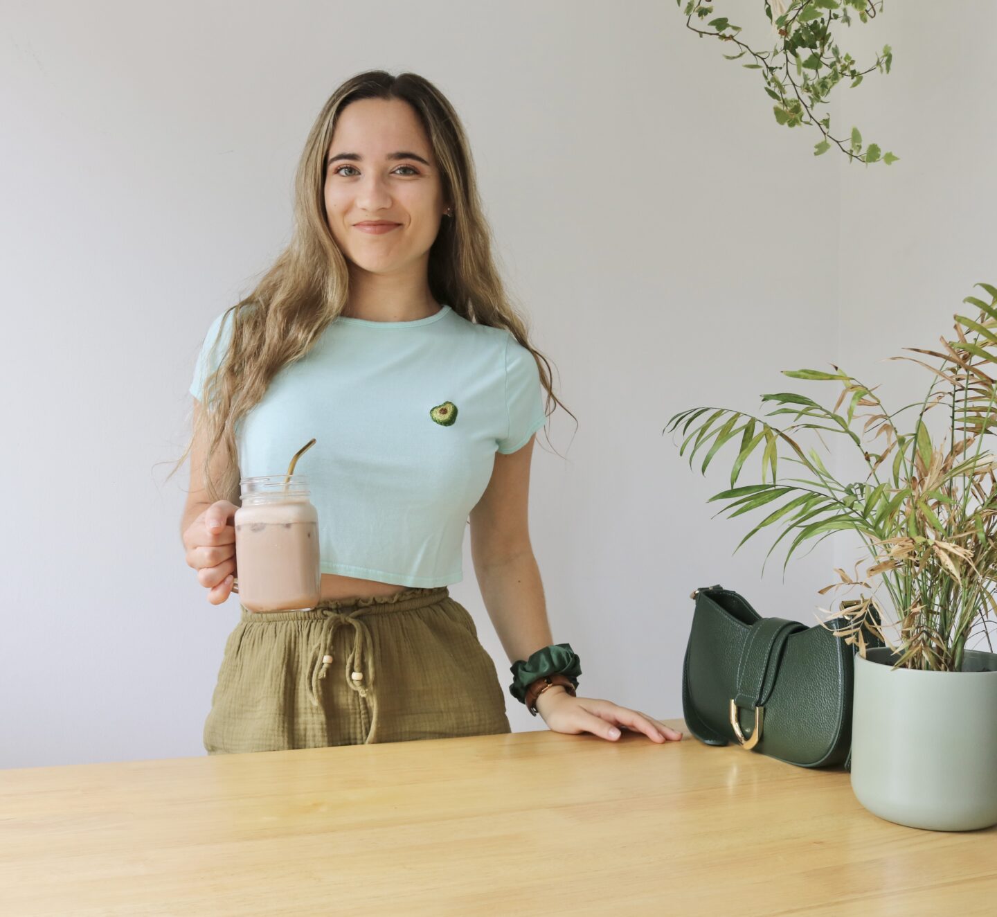 A brunette girl stood up holding an iced coffee with a plant next to her wearing a blue/green crop top hand embroidered with an avocado
