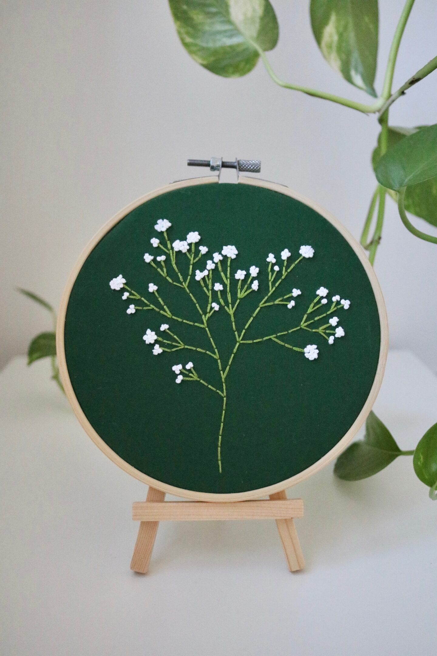 A 6 inch hoop with dark green fabric hand embroidered with a Gypsophila flower. The hoop is displayed in a wooden stand