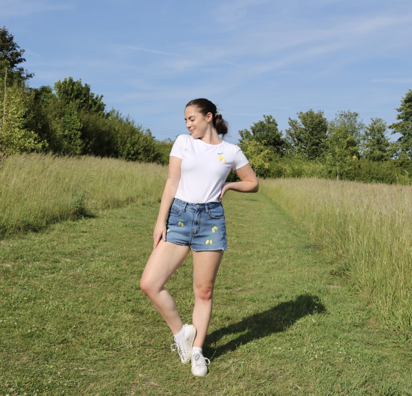 A brunette girl standing in a field on a sunny day wearing a white t-shirt hand embroidered with a pair of lemons and blue shorts hand embroidered with little lemons