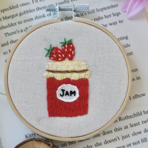 A close up of a hand-embroidered 3-inch hoop featuring a red strawberry jam jar with a golden fabric lid and two strawberries on top, stitched on natural cotton and framed in a wooden hoop, styled with pink and white flowers on an open book.