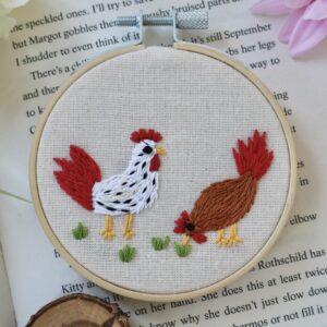 Hand-embroidered 3-inch hoop featuring two chickens, one white with black spots and one brown, stitched on natural cotton and framed in a wooden embroidery hoop, styled with flowers and an open book.