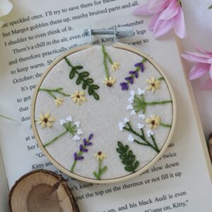 Hand-embroidered 3-inch hoop featuring white, yellow, and purple wildflowers on natural cotton fabric, framed in a wooden hoop and styled with soft pink and white flowers on an open book.