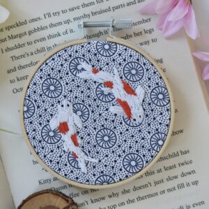 Hand-embroidered hoop featuring two orange and white koi fish on blue and white urchin-patterned Shweshwe fabric, framed in a wooden embroidery hoop.