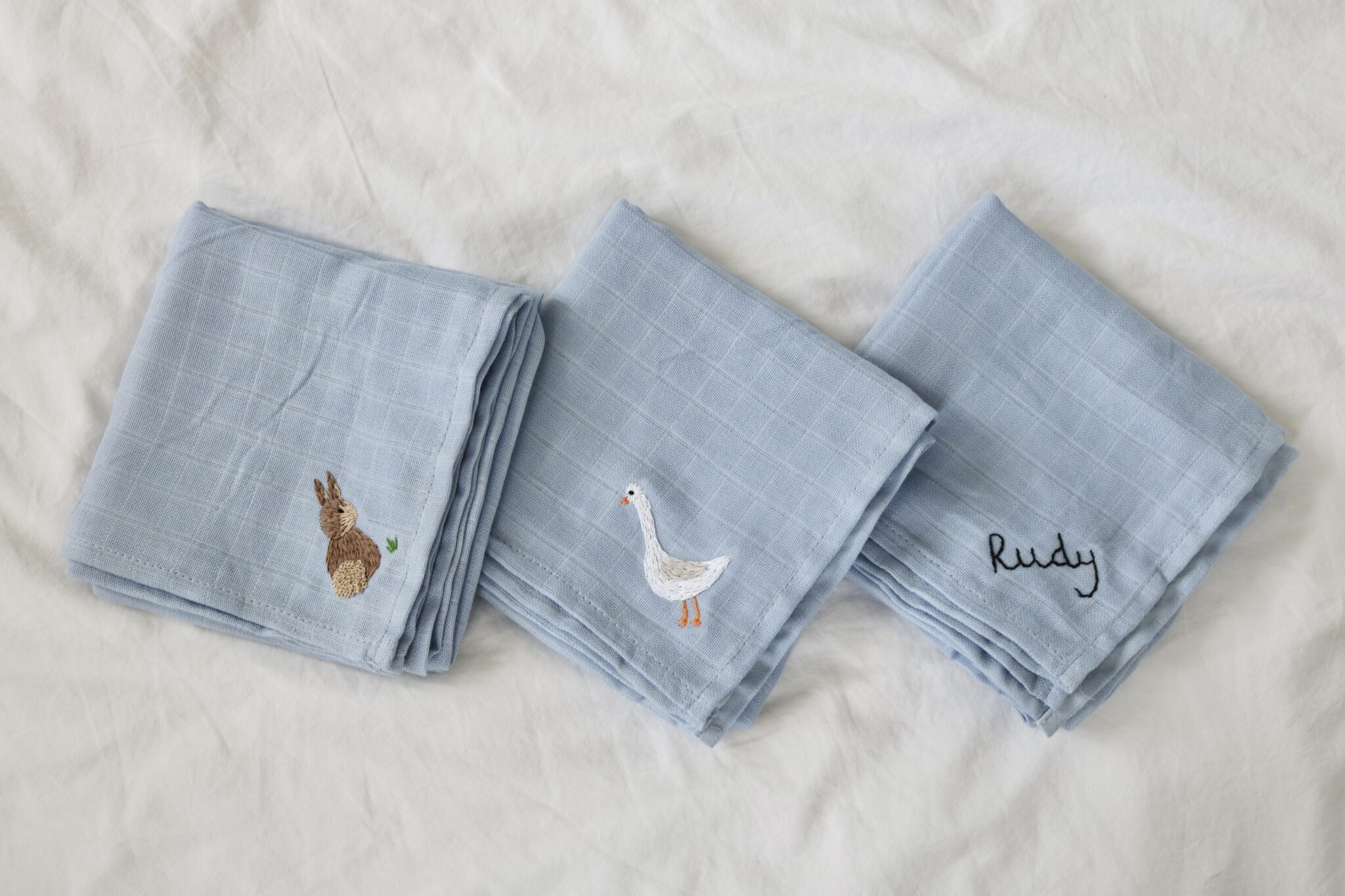 3 blue baby muslin cloths each hand embroidered with something different. One has a white goose, one has a brown bunny, and one has the name 'Rudy'