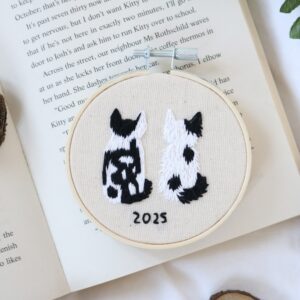 A 3 inch embroidery hoop hand embroidered with the back of two cats, both black and white, with the year 2025 embroidered below