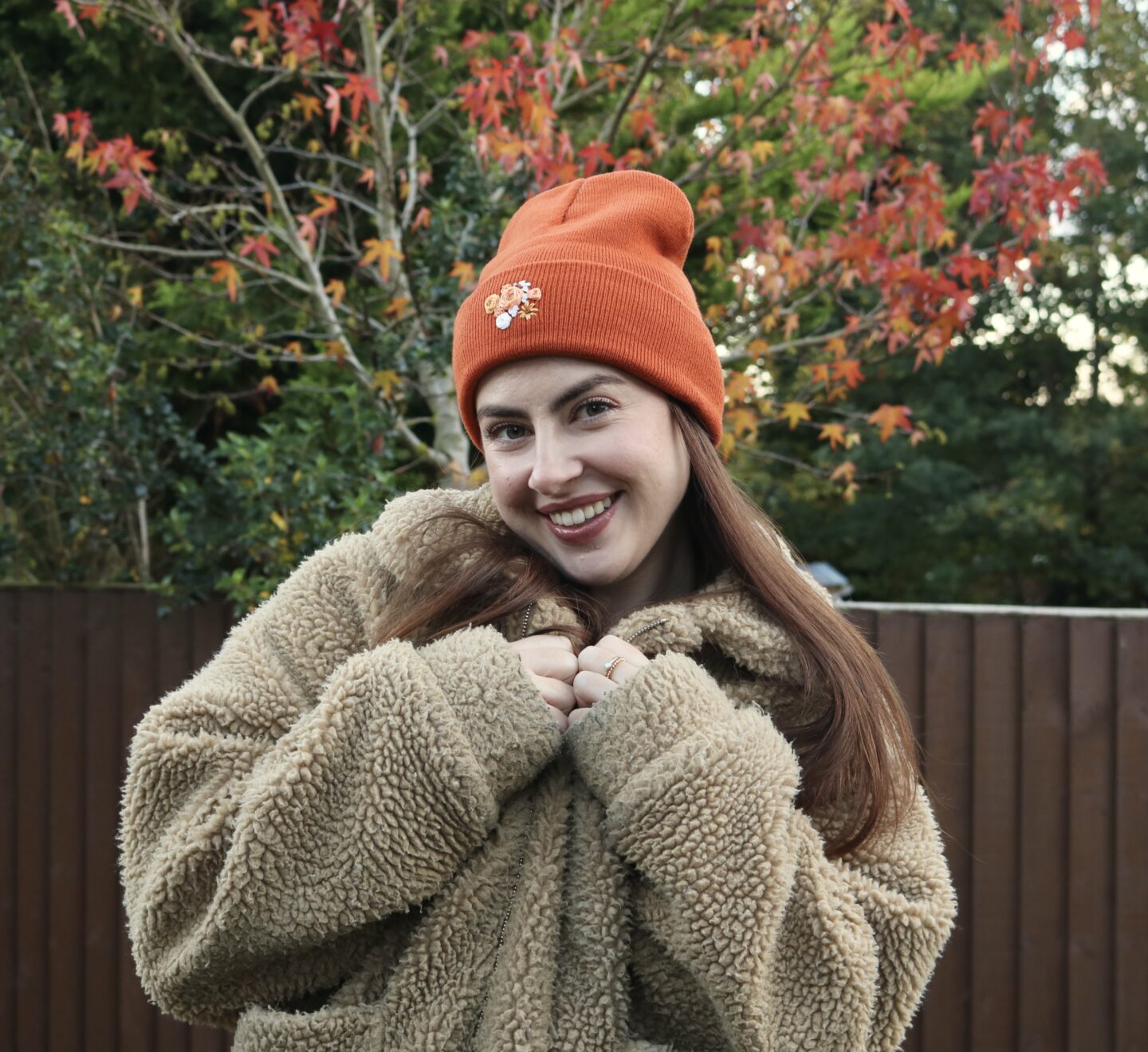 A brunette girl wearing fluffy brown coat smiling and wearing a rusty orange beanie hand embroidered with orange flowers
