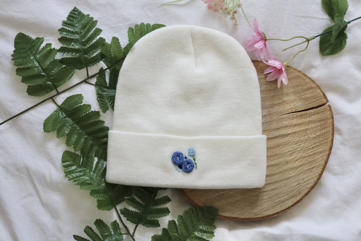A white knitted beanie hand embroidered with blue flowers featuring roses, cosmos, and a hydrangea