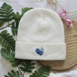 A white knitted beanie hand embroidered with blue flowers featuring roses, cosmos, and a hydrangea