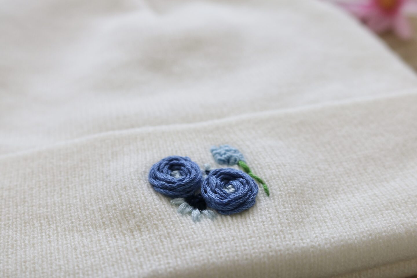 A close up of blue floral embroidery on a white knitted beanie hat. The floral embroidery features roses, cosmos, and a hydrangea