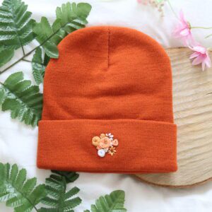 A rusted orange knitted beanie hand embroidered with orange hued flowers. Featuring roses, hydrangeas, and cosmos