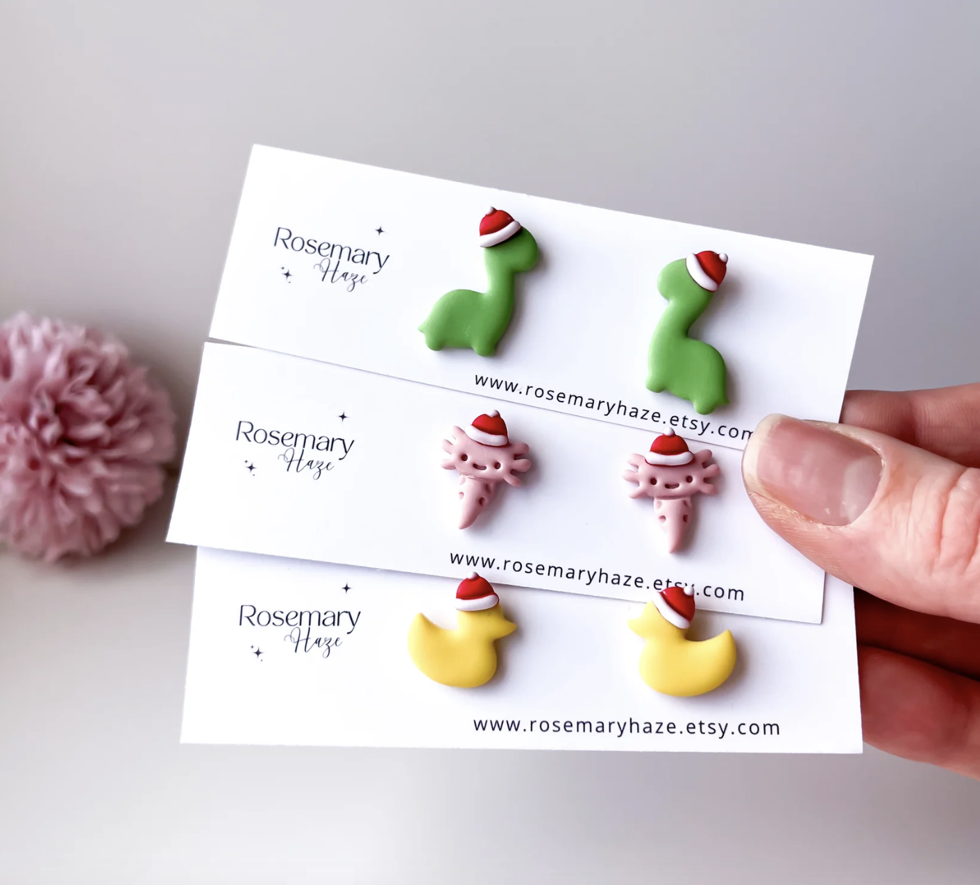 An image of three sets of earrings, one set of dinosaurs with a Christmas hat, one set of axolotl with Christmas hats, and one set of yellow ducks with Santa hats