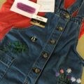 A flatlay of a denim pinafore dress hand embroidered with colourful flowers, bees, and a monstera leaf