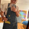 A brunette girl smiling and wearing a denim pinafore dress hand embroidered with a sunflower and bees
