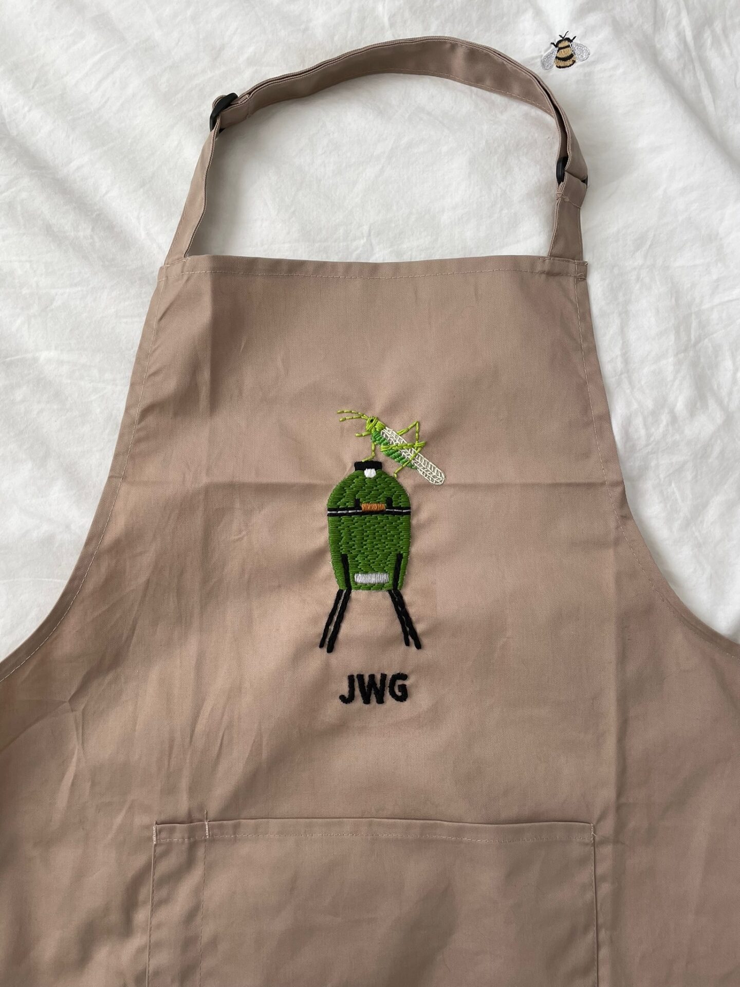 A beige apron hand embroidered with a green 'egg' bbq with a grasshopper on top and the initials DWG underneath