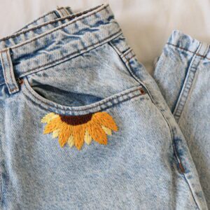 A pair of light wash blue mom jeans folded up showing the front left hand pocket embroidered with half a sunflower
