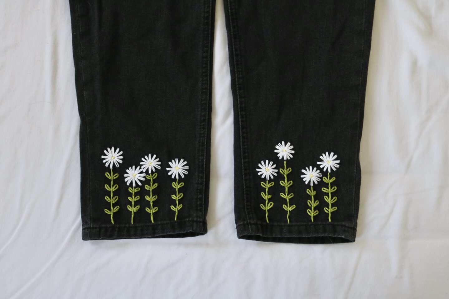 The two legs and cuffs of a pair of black denim jeans hand embroidered with four white daisies growing up each cuff