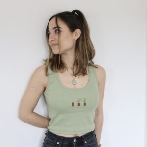 A brunette girl wearing black jeans and a soft green tank top hand embroidered with three cacti in the centre of the top