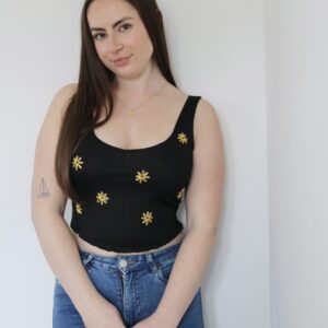 A brunette girl standing wearing a black crop top hand embroidered with sunflowers scattered across, paired with blue skinny jeans
