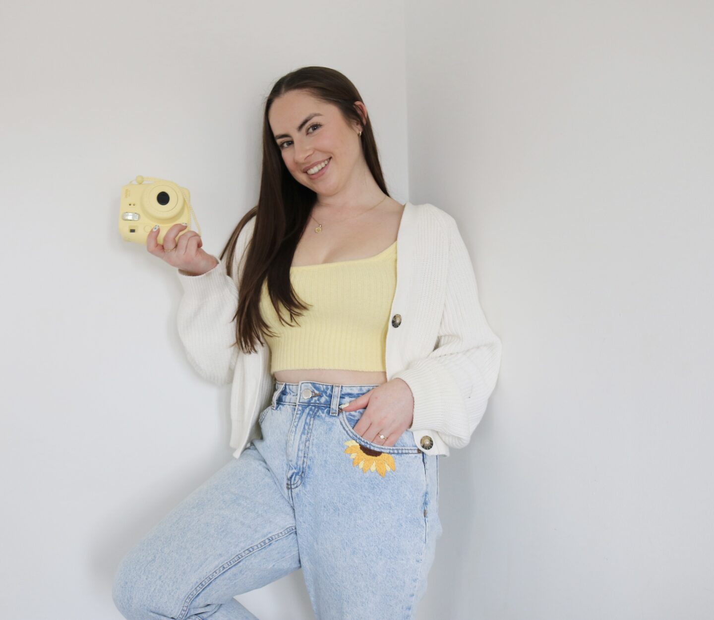 A brunette girl wearing a yellow knitted top and light blue mom jeans hand embroidered with a sunflower by the pocket