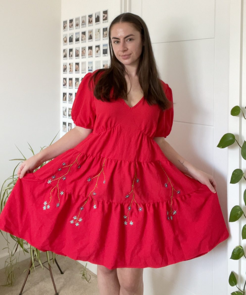A brunette girl wearing a red dress hand embroidered with blue forget me not flower vines on the skirt
