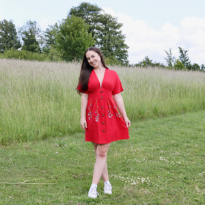 A brunette girl wearing a red button up dress hand embroidered with blue forget me not flower vines on the skirt