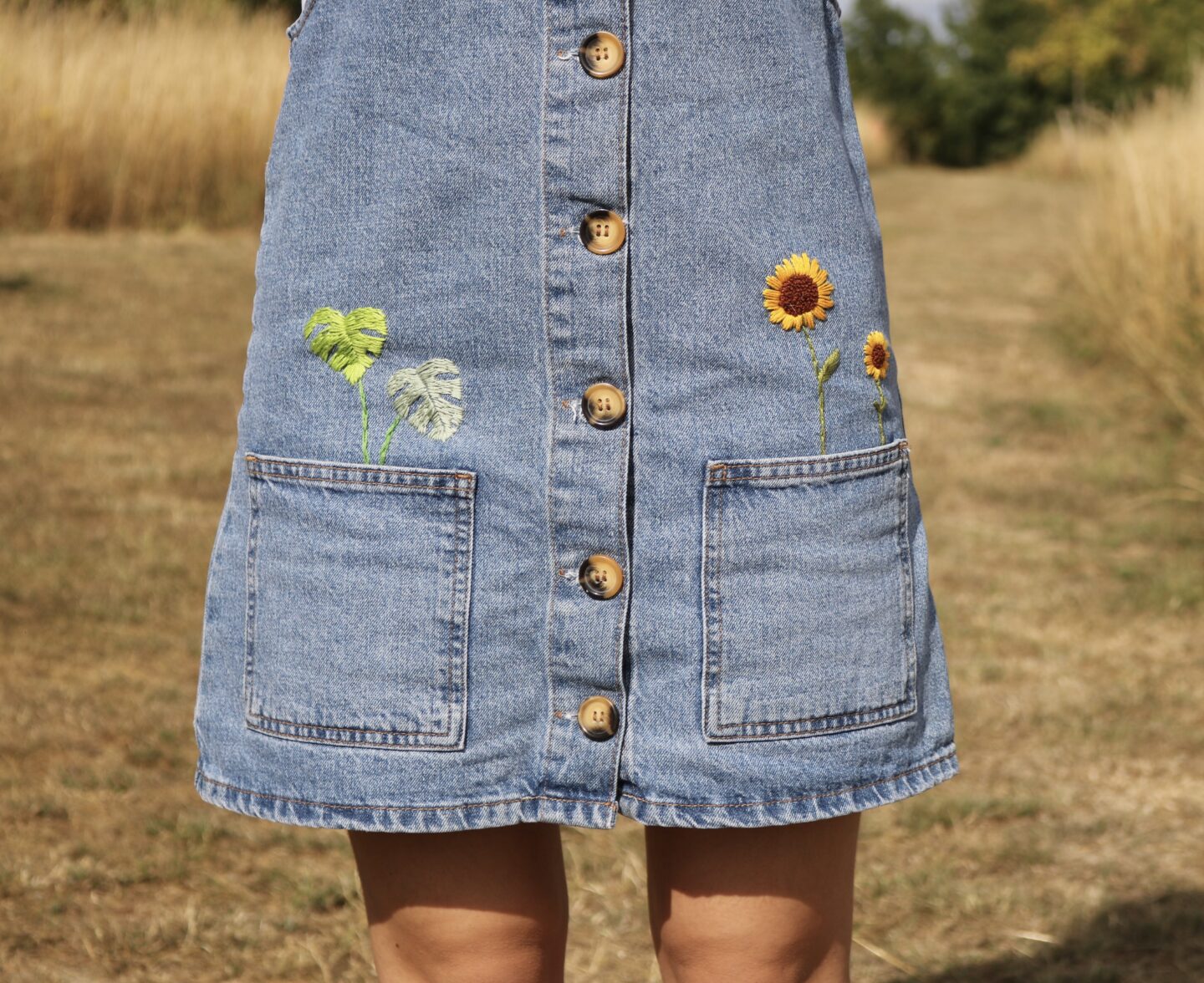 A blue denim pinafore hand embroidered with sunflowers and monstera leaves coming out the pockets