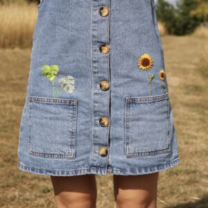 A blue denim pinafore hand embroidered with sunflowers and monstera leaves coming out the pockets